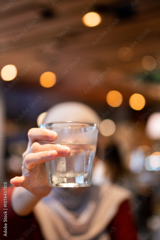 A person holds a glass of fresh water in a cozy cafe setting with blurred background. The warm, ambient lighting creates a relaxing and inviting atmosphere for hydration and reflection. A person holds a glass of fresh water in a cozy cafe setting with blurred background. The warm, ambient lighting creates a relaxing and inviting atmosphere for hydration and reflection.
