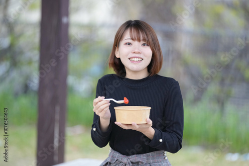 A young woman in her 20s with short hair wearing a black knitted long-sleeved shirt is sitting in a pavilion in an empty park and eating fruits such as dragon fruit, pineapple, and strawberries.