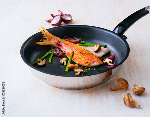 Freshly fried Red Mullet With Vegetables Sizzling In A Skillet. A vibrant red fish is perfectly cooked in a skillet alongside fresh asparagus, mushrooms, and onions.