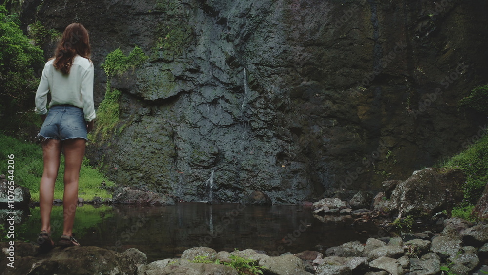 Obraz premium A woman stands on rock waterfall in French Polynesia. The tropical waterfall flows in a small pool, surrounded by lush greenery and volcanic rock. Peace and tranquility, showcasing nature beauty