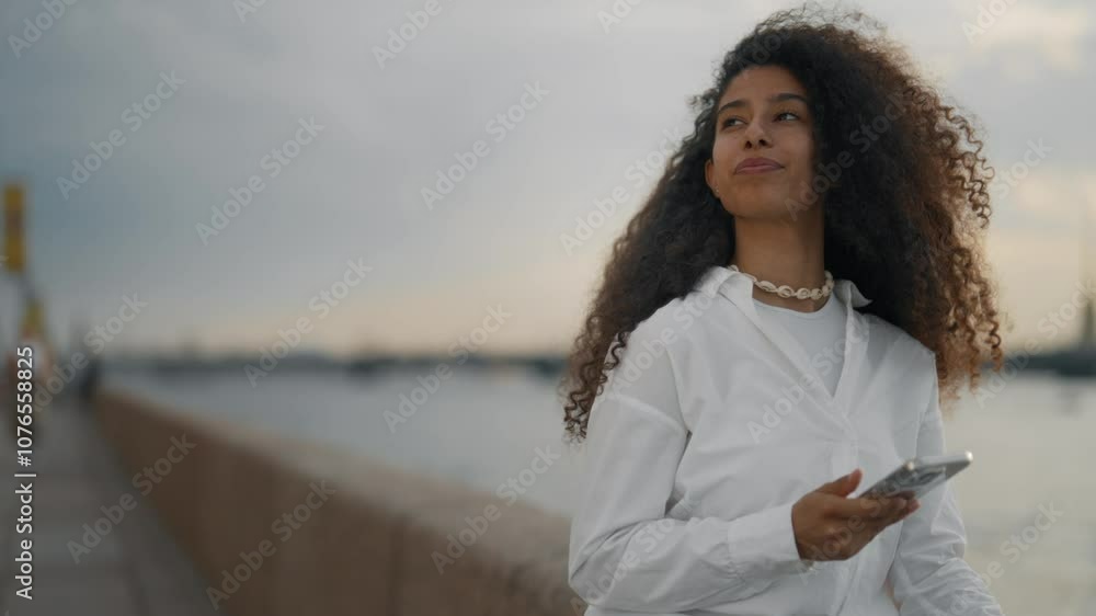Beautiful mixed race woman with modern smartphone on city embankment, portrait. Happy and joyful female city dweller relaxing in sunset, summer vacation in city, positivity and good mood, medium shot Beautiful mixed race woman with modern smartphone on city embankment, portrait. Happy and joyful female city dweller relaxing in sunset, summer vacation in city, positivity and good mood, medium shot