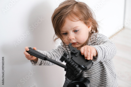  Toddler playing with a camera tripod in a bright room. A young child with brown hair explores a camera tripod in a bright room. Wearing a striped sweater, the child looks focused, with a serious expr photo