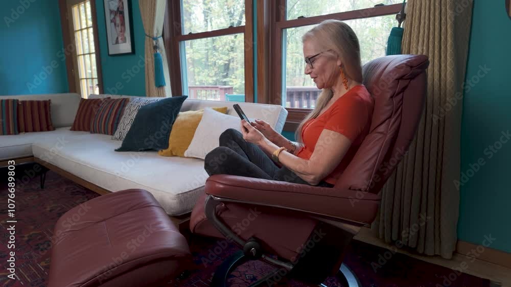 Excited mature woman in an orange shirt sits in a red leather recliner, using a smartphone. She is smiling and reading good news on the phone. Excited mature woman in an orange shirt sits in a red leather recliner, using a smartphone. She is smiling and reading good news on the phone.