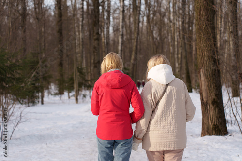 Two women walking together on a snowy path surrounded by bare trees in a winter forest, seen from behind.