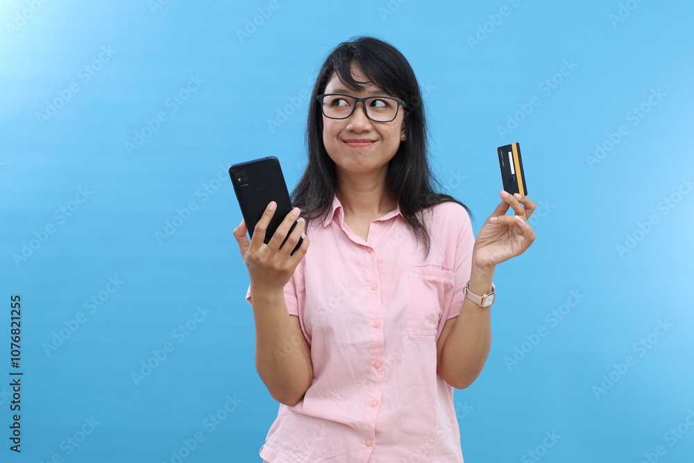 Smiling asian woman, lady in suit thinking, holding smartphone and credit card, plan to buy something online, shopping with mobile phone, blue background Smiling asian woman, lady in suit thinking, holding smartphone and credit card, plan to buy something online, shopping with mobile phone, blue background