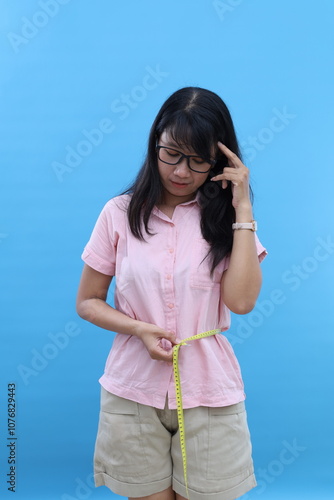 Young sad confused woman wears pink clothes hold measuring tape on waist spread hand isolated on blue background. Proper nutrition healthy fast food unhealthy choice concept