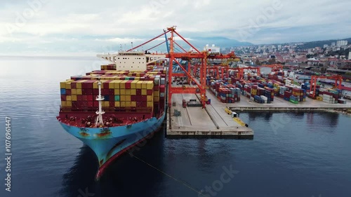 Process of loading, unloading ship with containers by cranes in logistics park. Aerial view of huge container ship, moored in cargo port in mountains. Global cargo transportation. Delivery of goods.