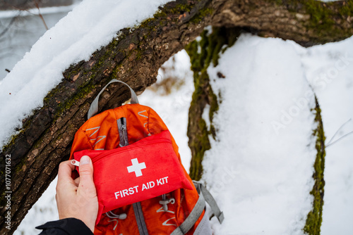 A person is standing in the snow, holding a first aid kit in one hand, and next to them, there is a backpack resting on the icy ground, prepared for any winter activities ahead photo