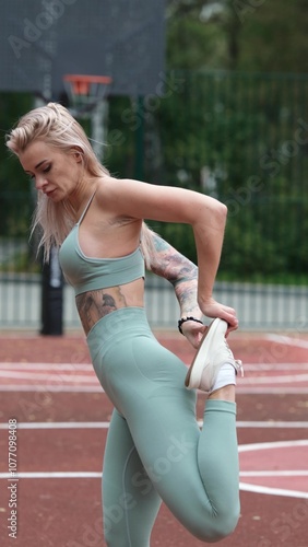 Sporty happy young blonde woman in sportswear doing leg stretching exercises standing on basketball court outside in the morning. Genuine candid photo photo