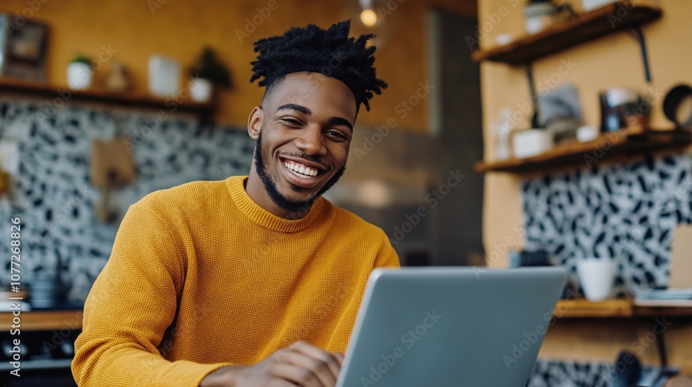 Fototapeta premium Smiling man in a yellow sweater using a laptop in a modern kitchen with patterned backsplash and wooden shelves.