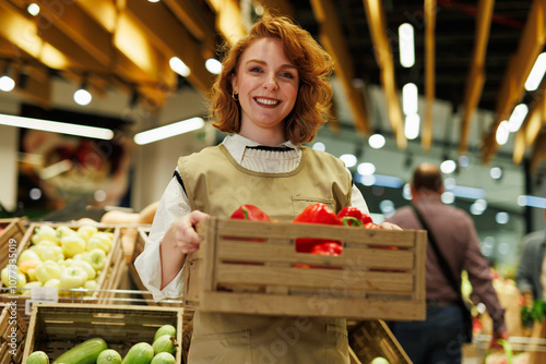 Photos Smiling greengrocer holding crate of red bell peppers in supermarket