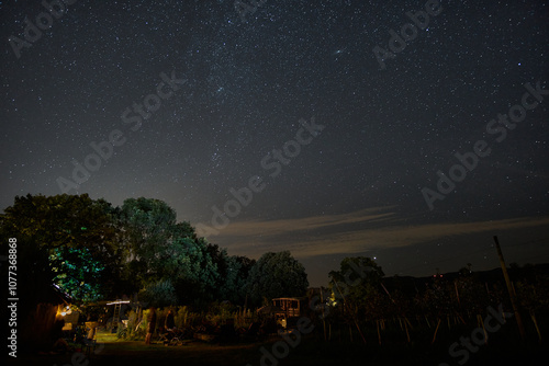 Starry night sky over a garden with trees and a distant building in a quiet countryside setting
