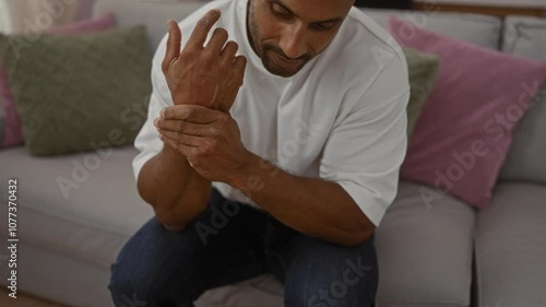 Young man indoors sitting on couch holding wrist in living room with cushions in casual attire