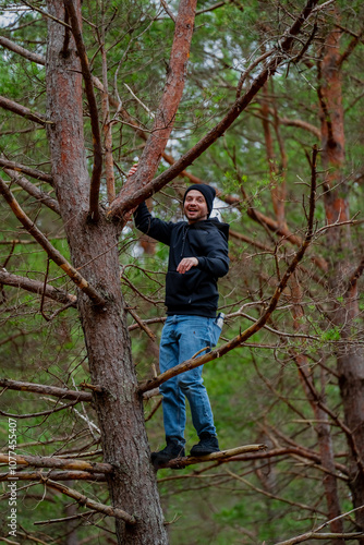 Man in casual clothing jeans and black hoodie, climbing a high pine tree as leisure activity in stroll in woods having fun