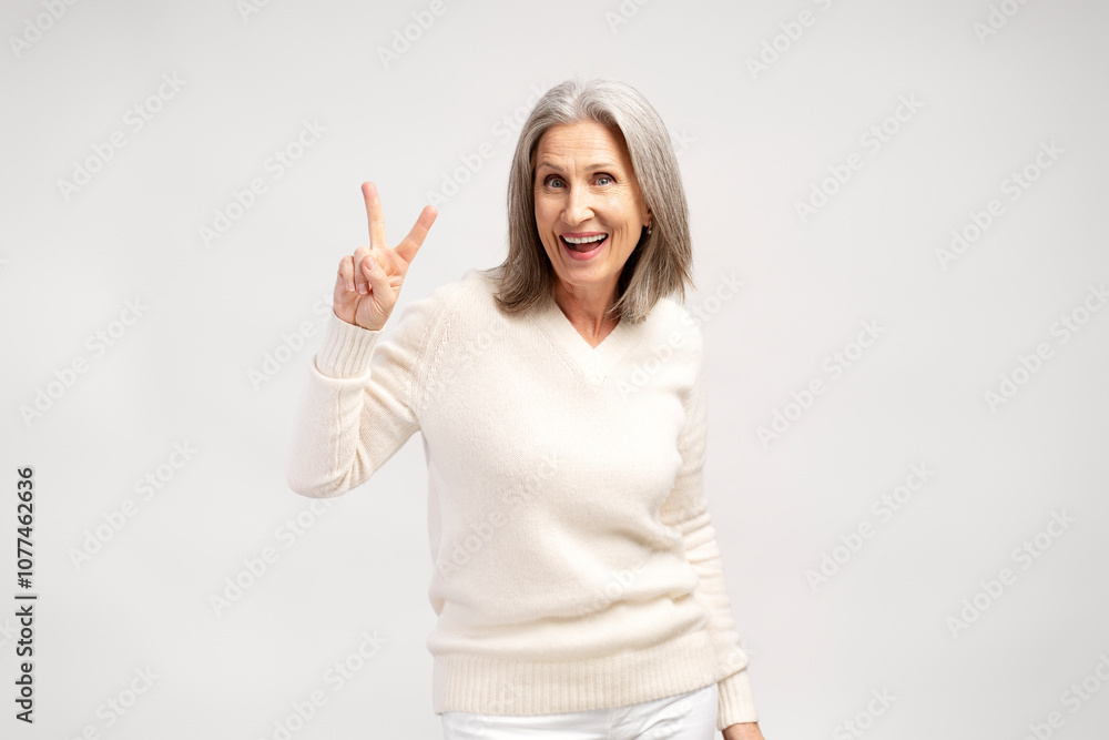Smiling, excited senior gray haired woman showing peace gesture, posing isolated on white background