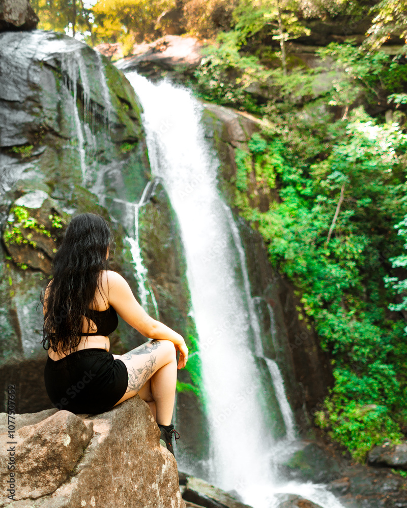 Woman sitting on rock in front of waterfall in southern United States with bright green leaves on summer afternoon Woman sitting on rock in front of waterfall in southern United States with bright green leaves on summer afternoon