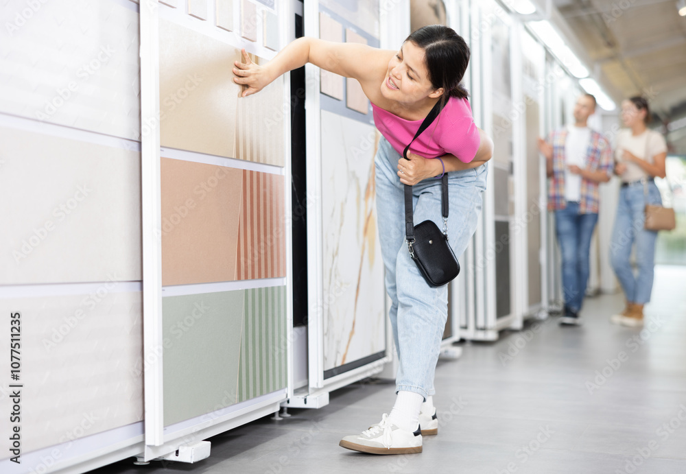 Interested young Asian woman examining technical stone tile samples displayed on sliding system exhibit stand in home improvement store, considering colors and textures for renovation project.. Interested young Asian woman examining technical stone tile samples displayed on sliding system exhibit stand in home improvement store, considering colors and textures for renovation project..