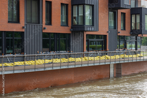 Flood protection measures with yellow sandbags and plastic sheeting safeguard a modern brick building along the riverside. Defense against high water levels and flood damage in an urban environment photo