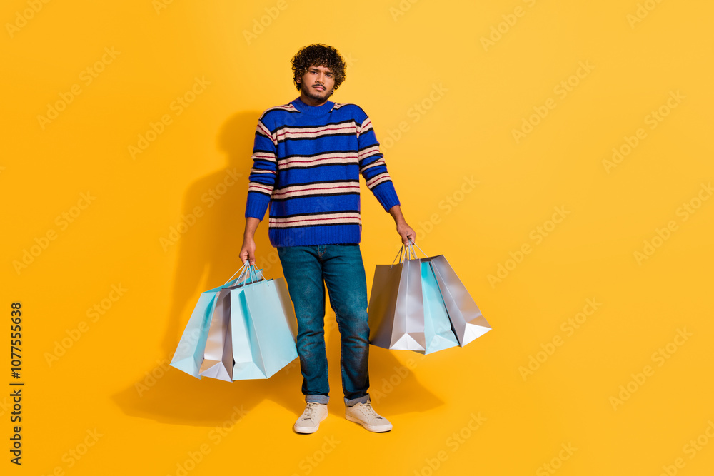 Full size photo of pretty young male hold shopping bags depressed wear trendy blue striped outfit isolated on yellow color background Full size photo of pretty young male hold shopping bags depressed wear trendy blue striped outfit isolated on yellow color background