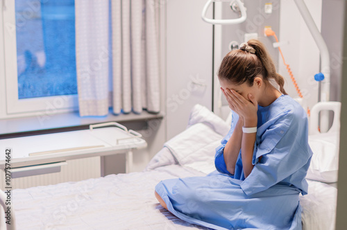 young woman in a blue hospital gown sits alone on a bed in a private room in a clinic, expressing her concern about the results of an examination, covering her face with both palms,