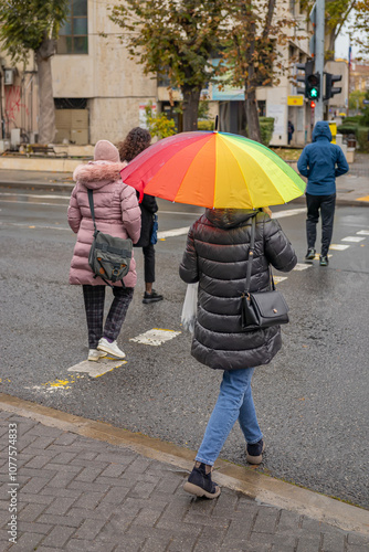 People walking on a rainy city street, woman with colorful rainbow umbrella, wearing warm coats