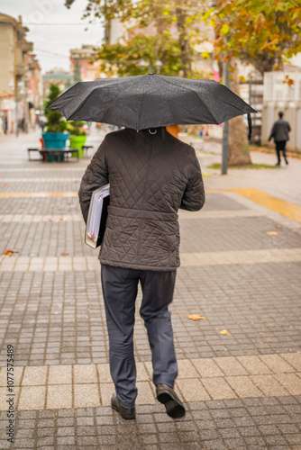 Rear view of man with a black umbrella walking down wet city street holding documents underarm photo