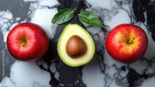 A halved avocado with a seed, two red apples and green leaves on a white and black marble surface. photo