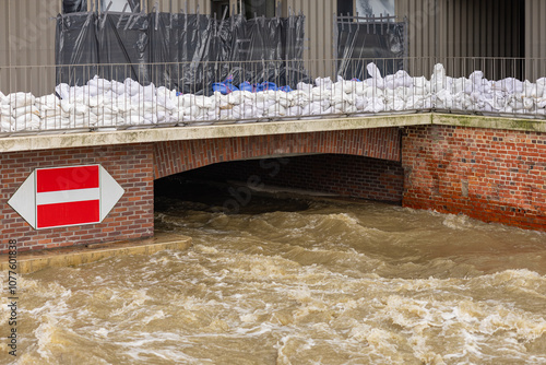 Flooded urban bridge with rapid water flow beneath and sandbags stacked for protection. High water levels submerge infrastructure, highlighting severe flooding and emergency response measures in city