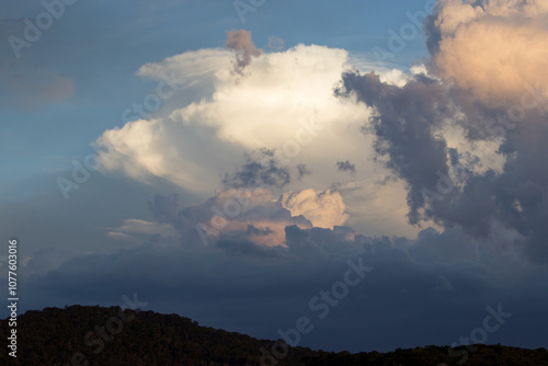 Dramatic sky with heavy rain clouds overlaying white clouds at dusk with stretch of forest covered mountains