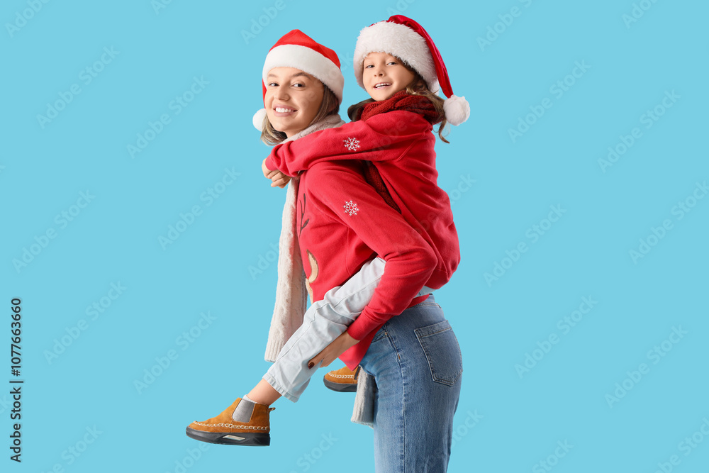 Fototapeta premium Beautiful young woman and her cute little daughter in Santa hats on blue background. Christmas celebration