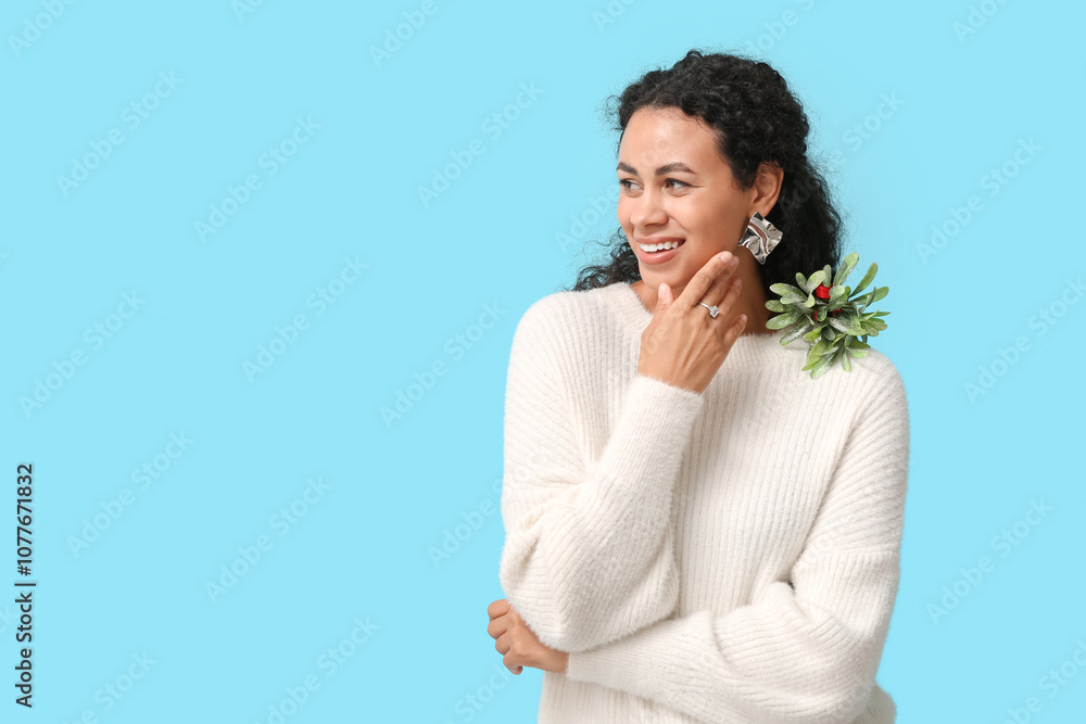 Beautiful young African-American woman in stylish earrings and ring with mistletoe branch on blue background. Christmas celebration Beautiful young African-American woman in stylish earrings and ring with mistletoe branch on blue background. Christmas celebration