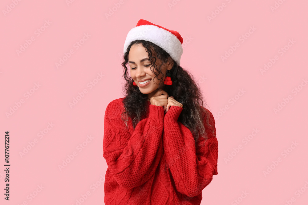 Beautiful young African-American woman in festive earrings and Santa hat on pink background. Christmas celebration