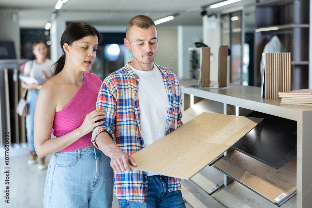 Obraz premium Interested young interracial couple discussing choice of eco-friendly wood grain laminate flooring, exploring samples displayed on rack in modern building hypermarket while planning home renovation..