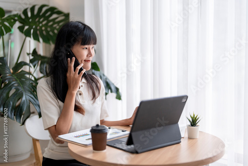 Asian businesswoman talking on phone and using tablet while working in modern office