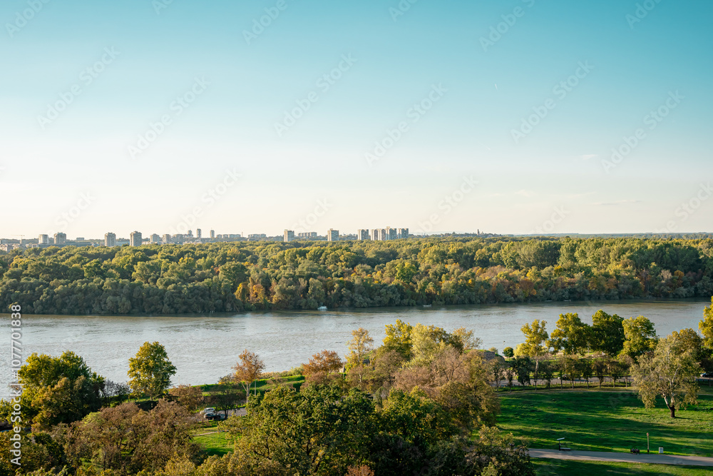 Fototapeta premium beautiful view of the city landscape during sunset, The Danube River meanders through a landscape where tall trees meet rolling hills in the background. The sky is partly cloudy, and the hills create