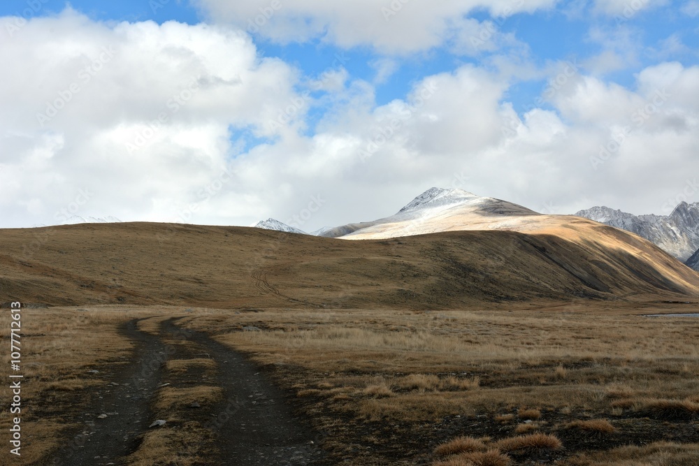 Bends of a field road climbing a high hill in the shadow of the clouds of the autumn sky against the backdrop of snow-capped mountain peaks. Bends of a field road climbing a high hill in the shadow of the clouds of the autumn sky against the backdrop of snow-capped mountain peaks.