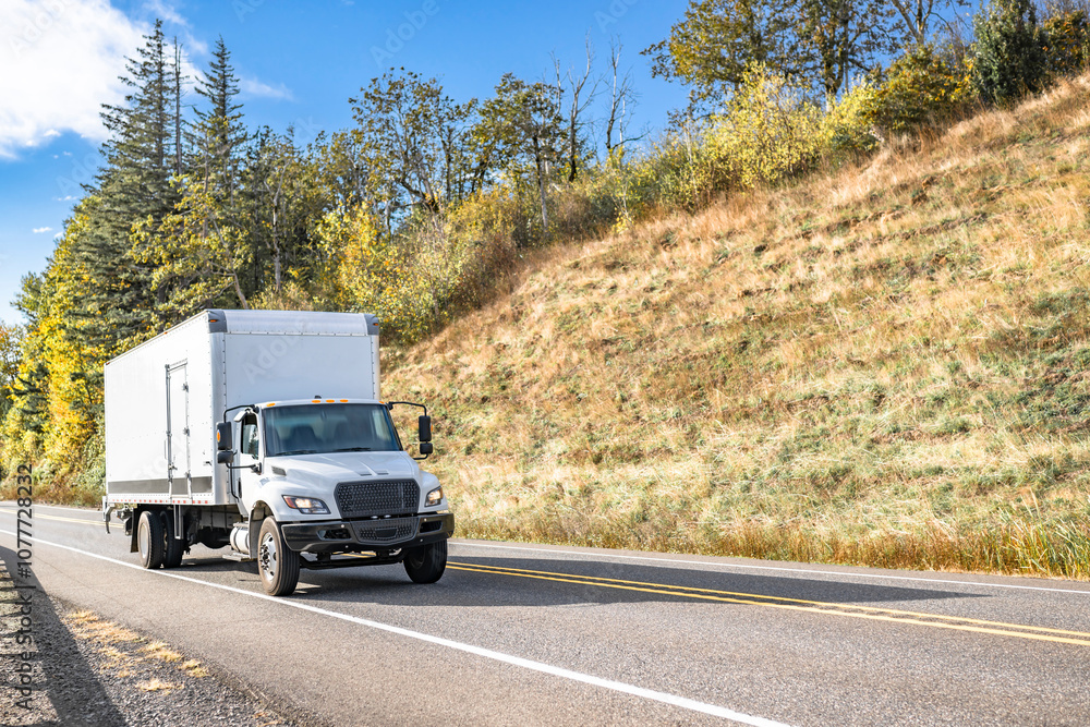 Obraz premium Compact white day cab rig semi truck transporting cargo in box trailer running on the narrow road between two hills with autumn trees