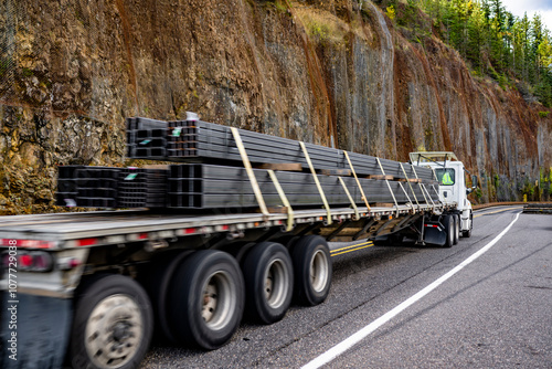 Day cab white big rig semi truck transporting heavy fastened metal cargo on flat bed semi trailer running on narrow mountain road with rock wall on the side photo