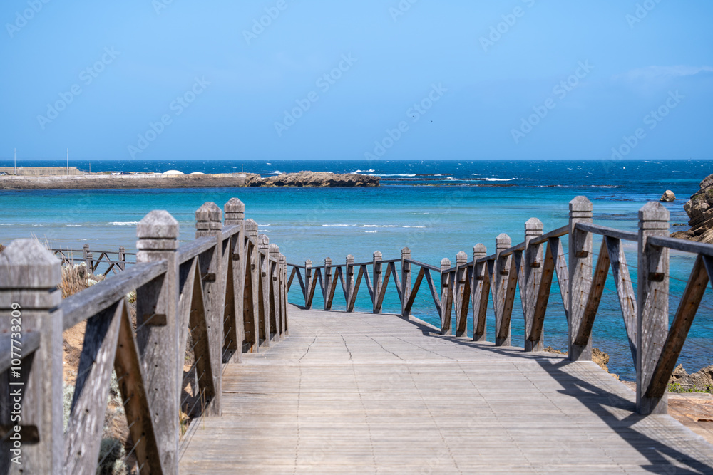 Wooden pedestrian boardwalk or walking trail leads to Merri Marine Sanctuary in Warrnambool, Australia with a beautiful turquoise-blue coastline with waves and rocky outcrops in the distance. Wooden pedestrian boardwalk or walking trail leads to Merri Marine Sanctuary in Warrnambool, Australia with a beautiful turquoise-blue coastline with waves and rocky outcrops in the distance.
