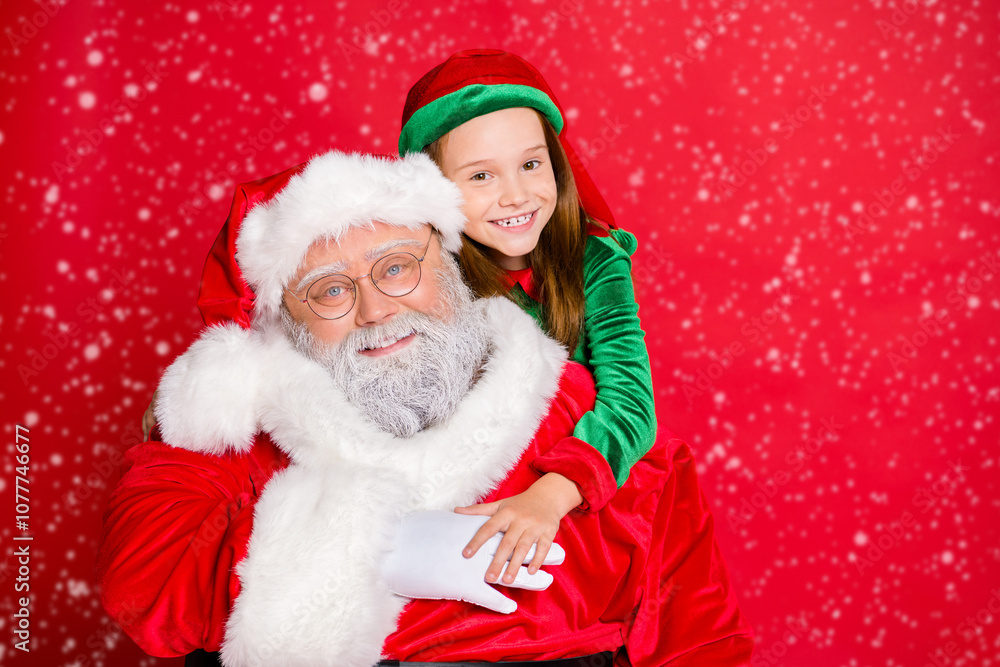 Close up photo of charming little elf helper in green hat headwear hugging piggyback her funny santa claus wearing eyewear eyeglasses isolated over red background Close up photo of charming little elf helper in green hat headwear hugging piggyback her funny santa claus wearing eyewear eyeglasses isolated over red background