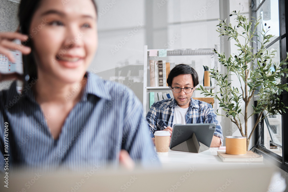 Fototapeta premium Asian Japanese male startup worker concentrates on working with digital tablet at desk near daylight window in casual small business office, online freelance jobs, and female colleague foreground.