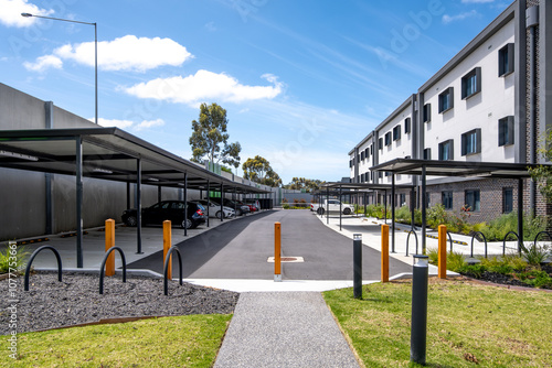 Modern apartment complex building, car park with covered parking bays or carports, and pedestrian walkways in a residential development. An organized parking infrastructure with bollards.