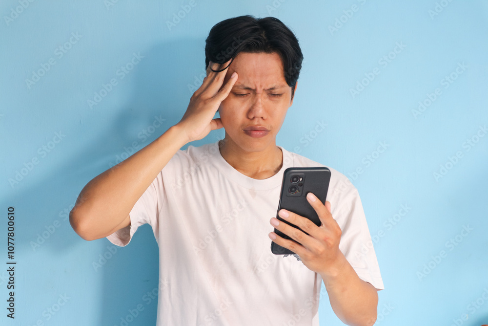 Young Asian man looking at mobile phone with sad expression while holding head Young Asian man looking at mobile phone with sad expression while holding head