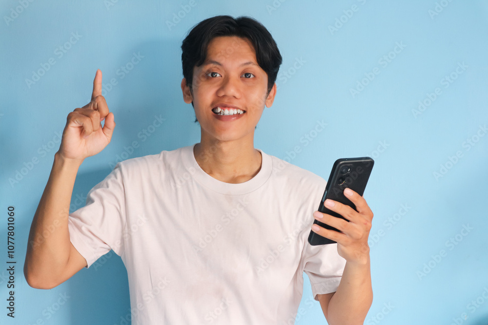 Asian young man holding at his mobile phone with happy expression while pointing finger up Asian young man holding at his mobile phone with happy expression while pointing finger up