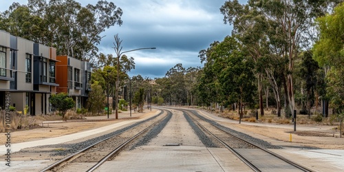 A quiet street with railway tracks bordered by trees and modern buildings under cloudy skies.