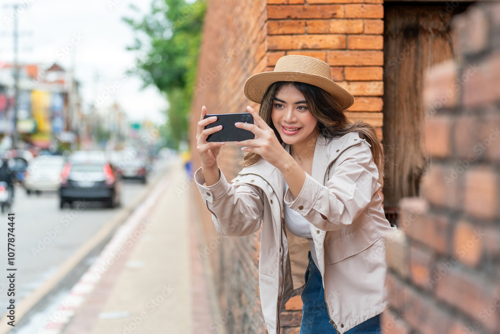 Fototapeta premium Asian Tourist Woman In Hat and Jacket Using Phone Beside A Brick Wall in Chiang Mai, Thailand