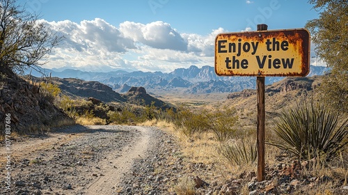 A weathered sign encourages travelers to pause and enjoy the magnificent views along a winding dirt road, surrounded by mountains and rolling hills under a bright sky