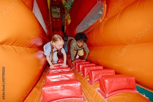 Cute little intercultural friends climbing soft orange leather tunnel with red stairs on playground while moving to steep slide