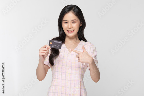 Cheerful Asian young woman confidently holds up credit card with bright smile against plain white background. Dressed in light stylish blouse exudes positivity and financial empowerment