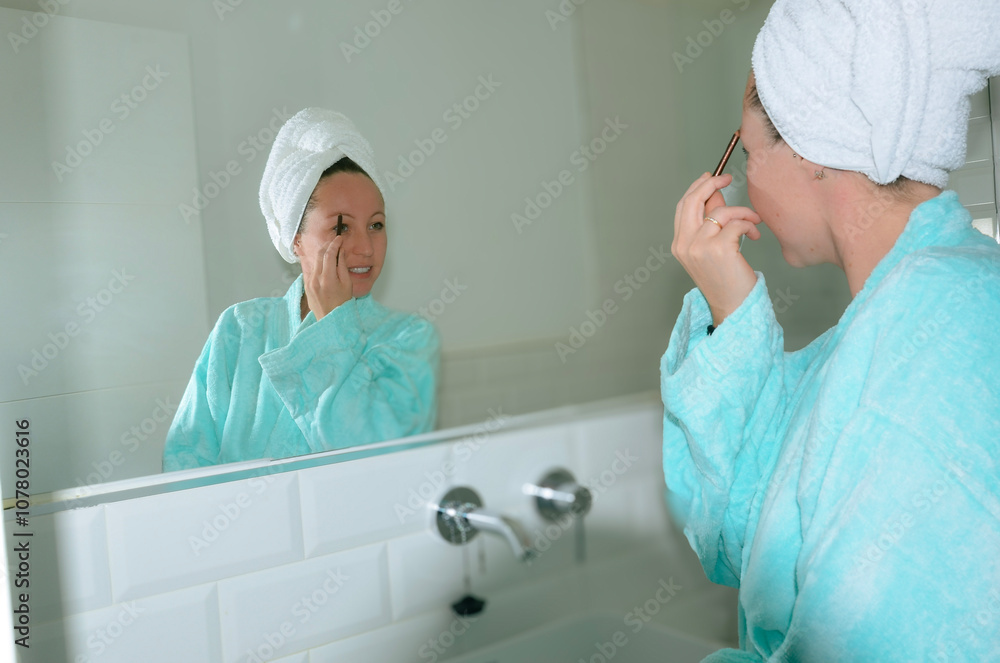 Obraz premium Woman applying makeup in bathroom mirror wearing bathrobe and towel turban